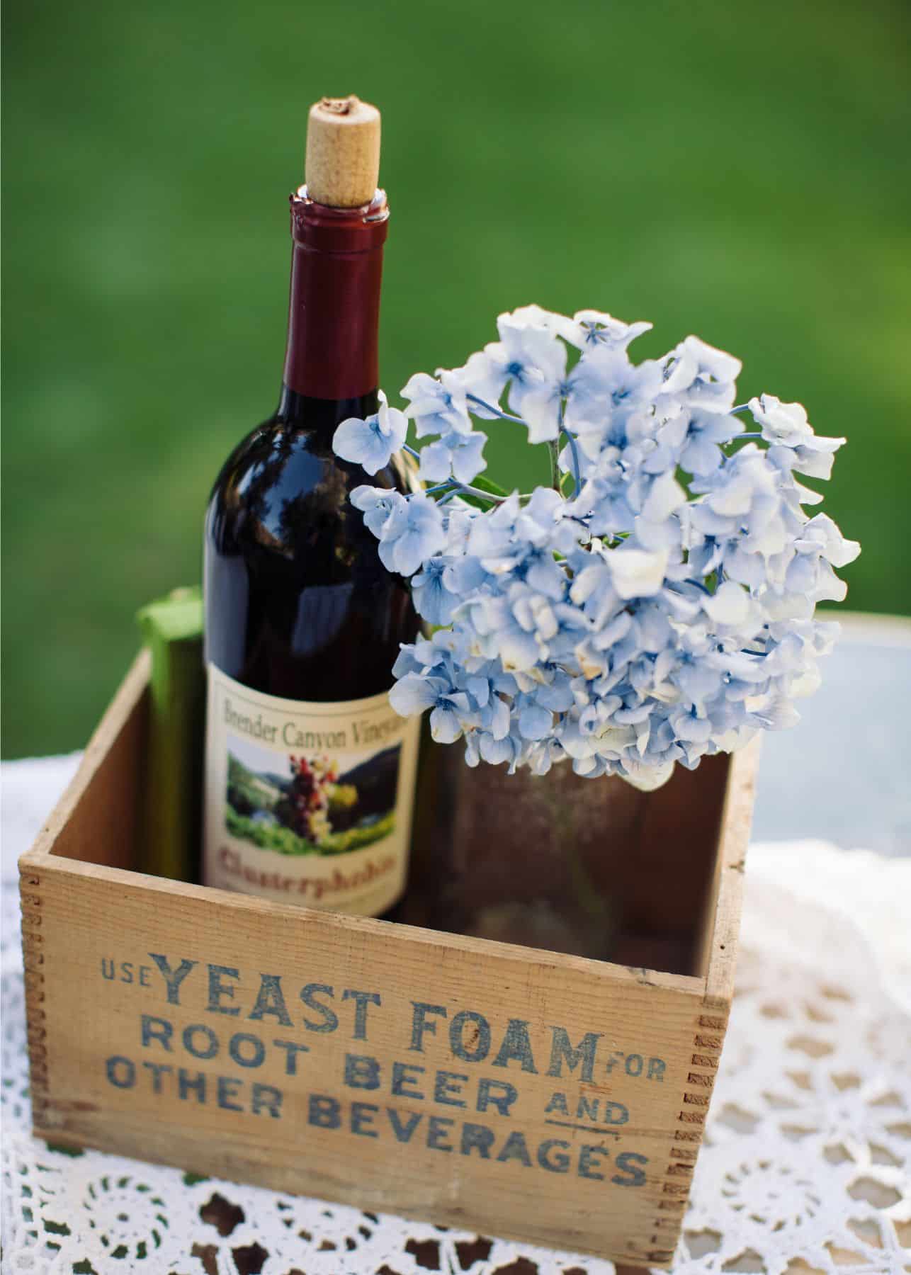 A rustic wooden box contains a bottle of wine and a bouquet of blue flowers.
