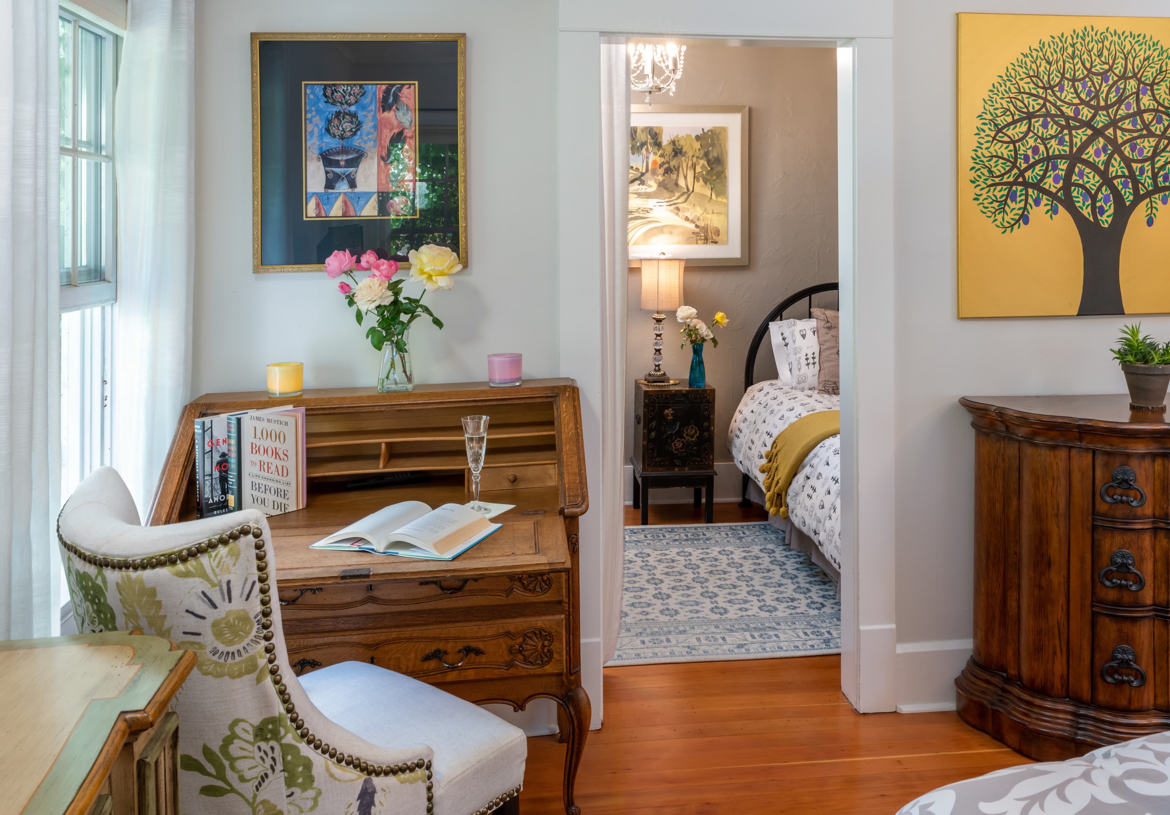 A cozy room featuring a vintage desk, a comfortable chair, and colorful artwork, leading to a softly lit bedroom.