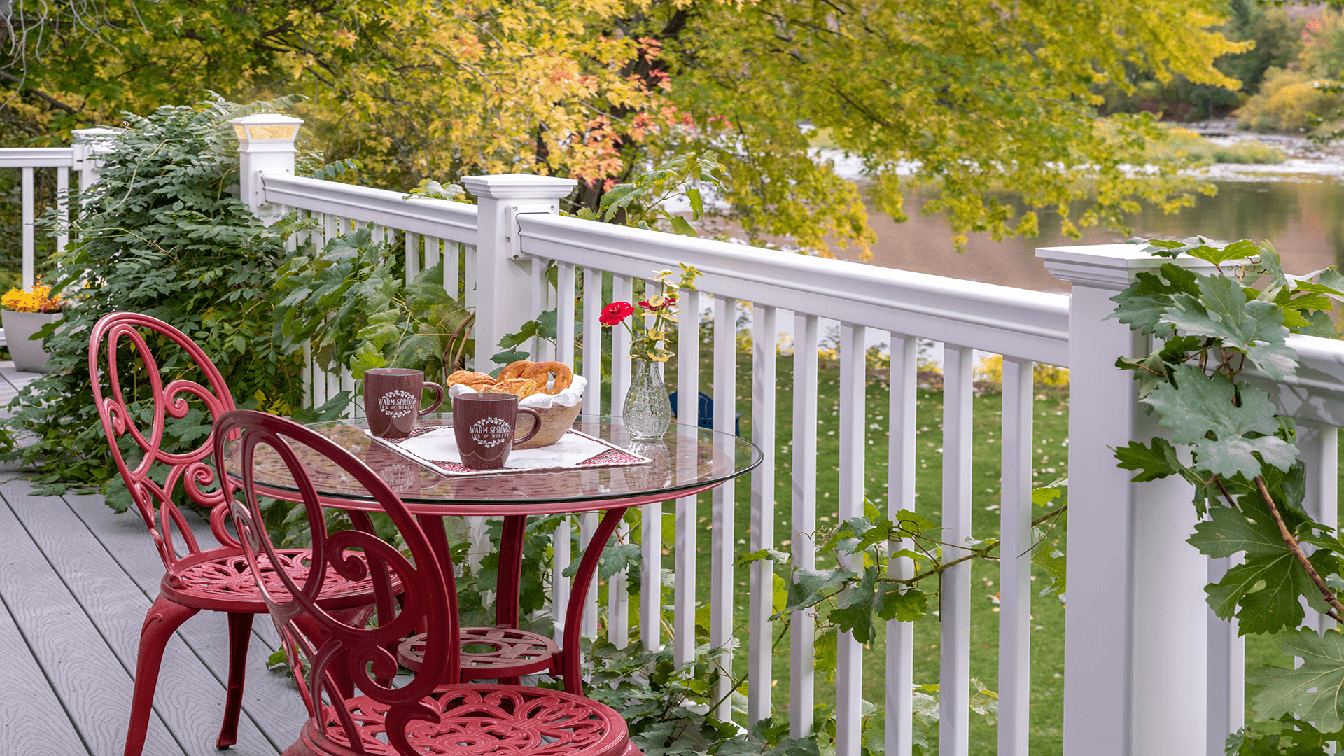 A quaint outdoor table set with coffee, pastries, and fresh flowers overlooks a serene river surrounded by autumn foliage.