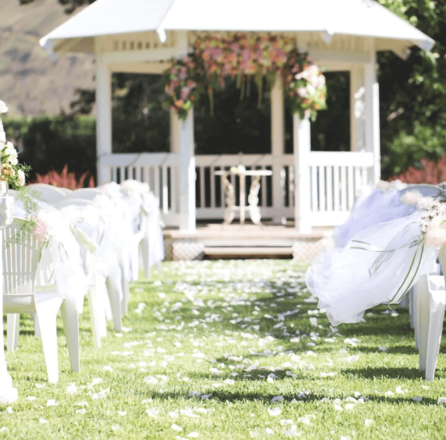 A picturesque outdoor wedding aisle lined with white chairs and flower petals leading to a decorated gazebo.