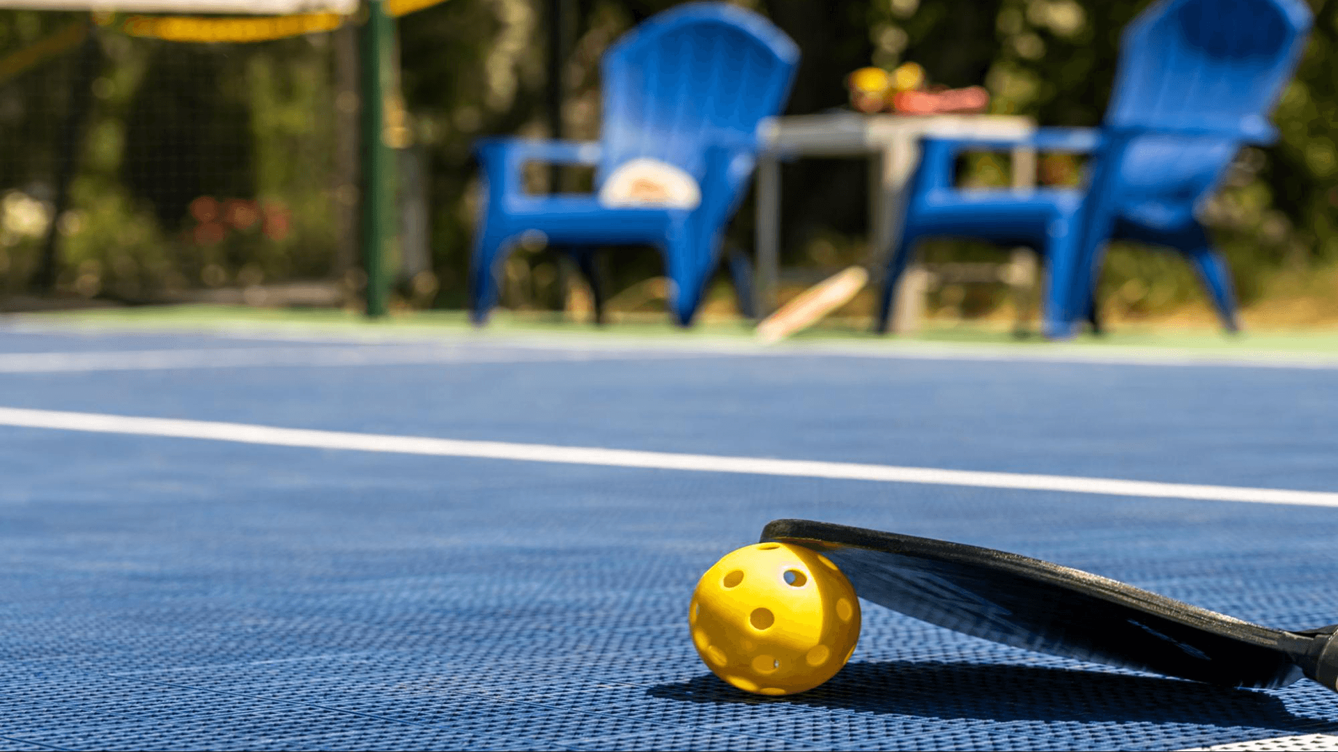 A yellow pickleball rests against a paddle on a blue court, with blue chairs in the background.