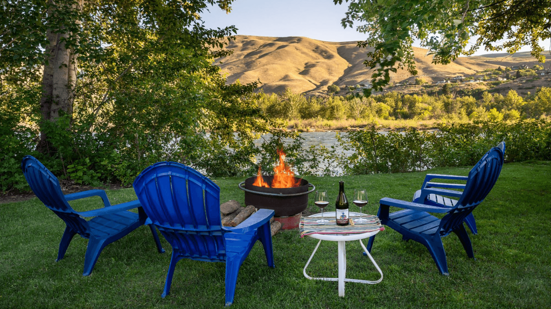 A cozy outdoor setting with blue chairs, a fire pit, and wine, overlooking a scenic river and hills.
