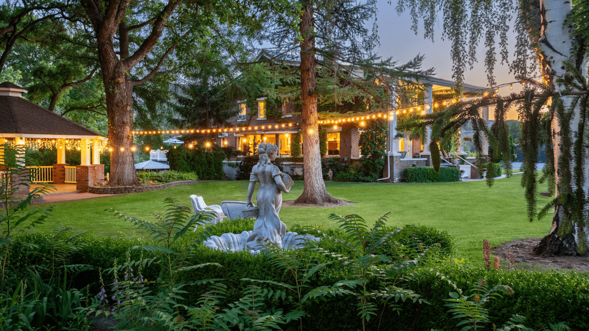A serene garden featuring a statue, illuminated string lights, and a charming house in the background.