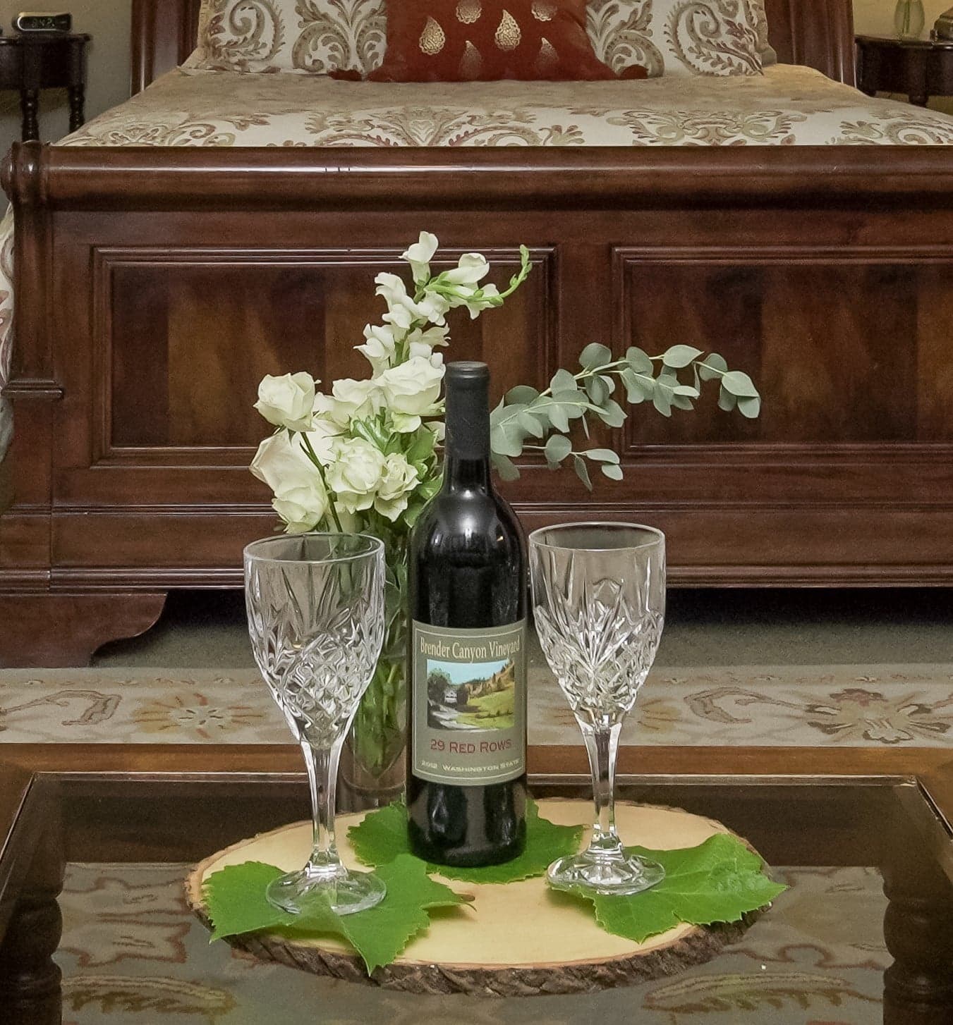A bottle of red wine and two crystal glasses on a wooden table, accompanied by a bouquet of white flowers.