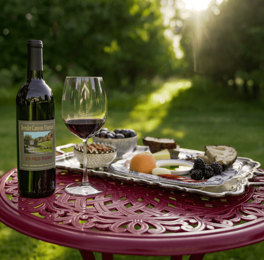 A bottle of red wine and a glass are accompanied by a platter of fruits, nuts, and bread on a table in a sunlit outdoor setting.